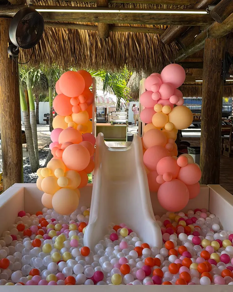 Ball pit with a slide framed by coral and peach balloon columns under a tiki hut